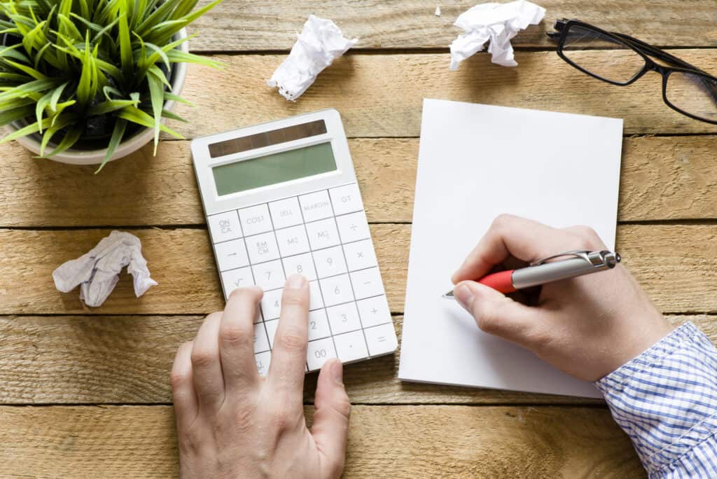 Man writing on notepad and using solar calculator on wooden desk, wondering, "Do solar calculators have batteries?"